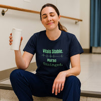 Real nurse in scrubs wearing “Vitals Stable. Nurse Unhinged.” t-shirt sitting in hospital break room, surrounded by nurse life clutter