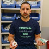 Real nurse in scrubs wearing “Vitals Stable. Nurse Unhinged.” t-shirt sitting in hospital break room, surrounded by nurse life clutter