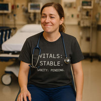 Nurse in ER wearing “Vitals: Stable. Sanity: Pending.” shirt, smiling tiredly with stethoscope around neck, seated in front of a hospital bed.