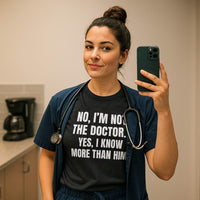 Female nurse in scrubs taking selfie, wearing "No, I’m Not The Doctor. Yes, I Know More Than Him." shirt, hospital setting.
