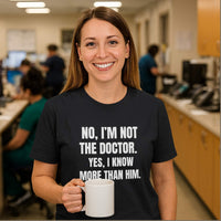Smiling nurse holding coffee mug, wearing "No, I’m Not The Doctor. Yes, I Know More Than Him." shirt, hospital nurse station.