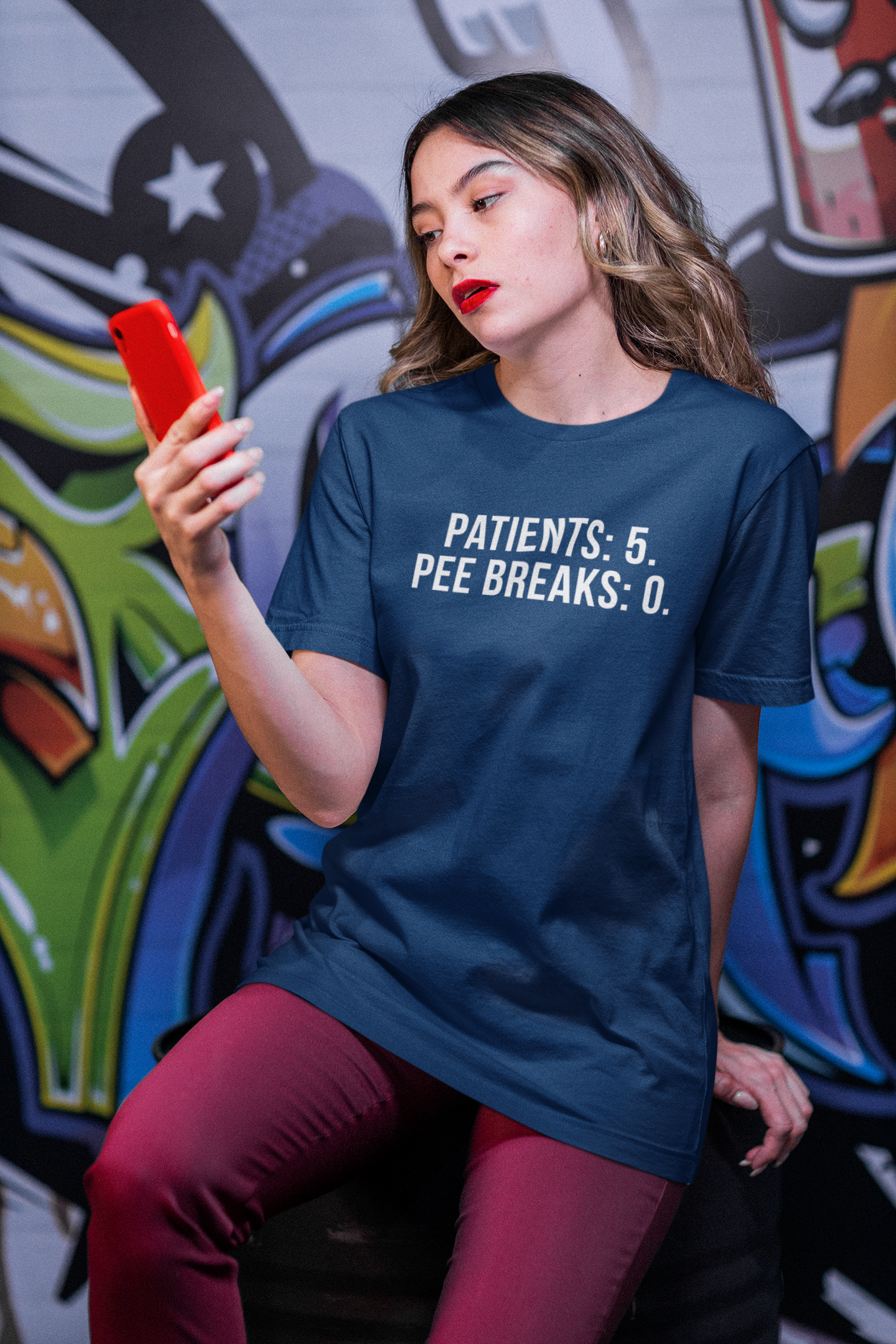 Nurse leaning against modern building wall, dressed in “Patients: 5. Pee Breaks: 0.” shirt.