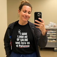Female nurse in scrubs taking selfie, wearing "I Gave 1,000 ml of Saline and Zero ml of Patience" shirt, hospital setting.

