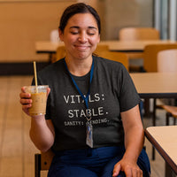 Nurse in scrubs grinning while wearing “Vitals Stable. Sanity Pending.” shirt in hospital hallway, actual shift energy, not stock
