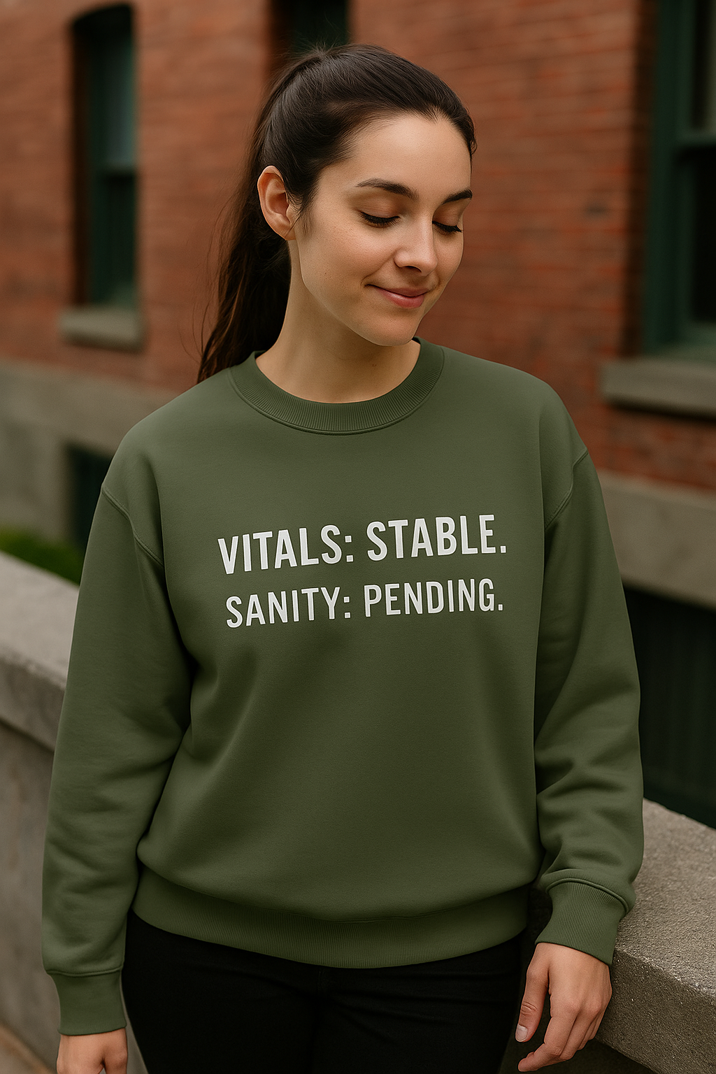 Female nurse in olive green crewneck sweatshirt with quote “Vitals: Stable. Sanity: Pending.” smiling outside hospital
