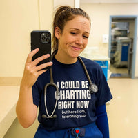 Female nurse taking selfie in scrubs, wearing "I Could Be Charting Right Now" shirt, clinical background.
