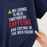 Close-up of shirt print: My License Is Held Together By Caffeine And Crying In The Med Room, warning triangle icon, colorful text on black fabric.
